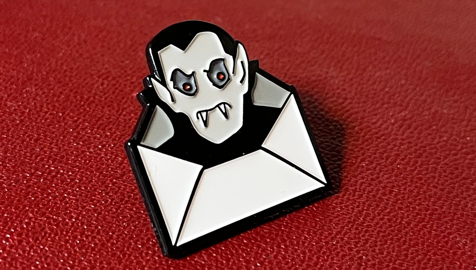 A Dracula Daily enamel pin featuring a stylized vampire face with fangs emerging from a white envelope, set against a textured red background.