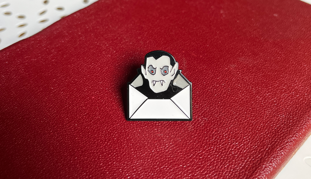 A small Dracula Daily pin shaped like a vampire face peeking out of an open envelope, placed on a textured red surface.