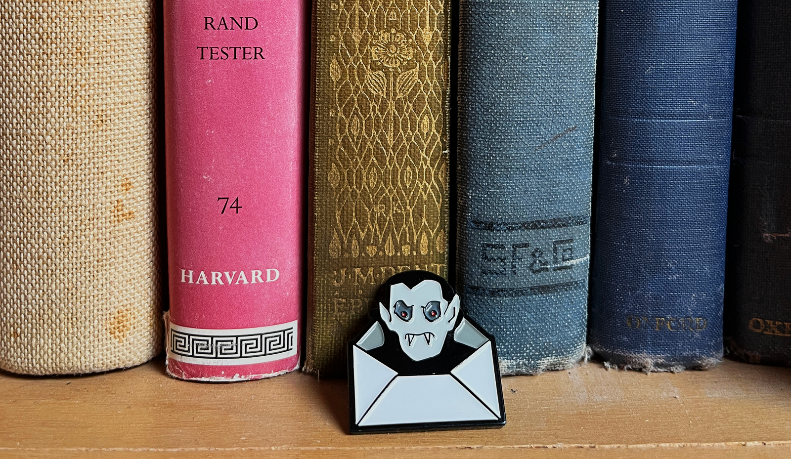 A Dracula Daily pin shaped like a vampire peeking out of an envelope, placed in front of a row of vintage hardcover books on a wooden shelf.