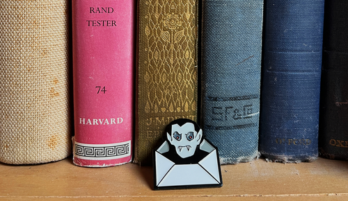 A Dracula Daily pin shaped like a vampire peeking out of an envelope, placed in front of a row of vintage hardcover books on a wooden shelf.