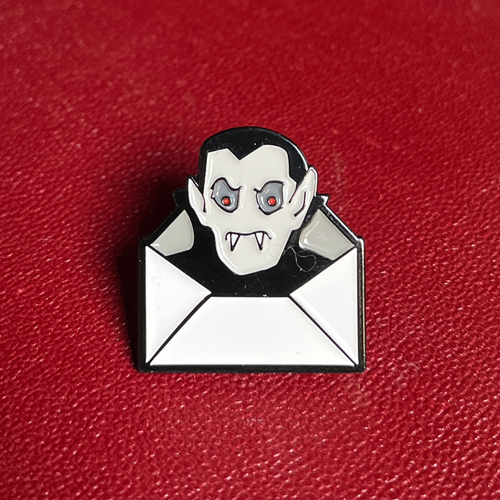 A Dracula Daily enamel pin with a white envelope, featuring a vampire face with black hair, pale skin, red eyes, and fangs, set against a textured red background.
